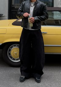 Svart skinnjakke over en grå langermet skjorte, kombinert med løse sorte bukser og svarte sko. Holder en gjennomsiktig flaske, står ved siden av en gul bil.