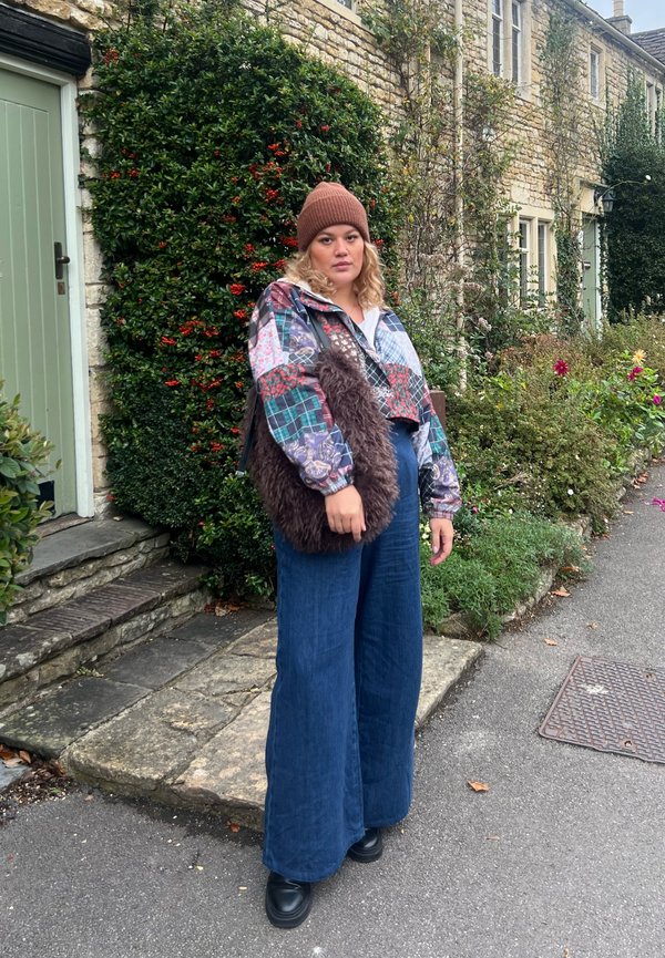 Chaqueta corta de múltiples patrones con un bolso de piel de pelo, pantalones anchos azules y un gorro de punto marrón. Rodeada de vegetación y un sendero de piedra.