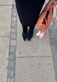 Czarne zamszowe botki za kostkę z ostrymi noskami i dekoracyjnym przeszyciem, w połączeniu z torbą ze splecionego różowego materiału i małym, puszystym breloczkiem.