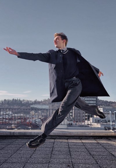 Abrigo negro con solapas anchas, suéter negro y pantalones grises usados por una persona en una pose dinámica contra un cielo despejado. Se ven zapatos negros.