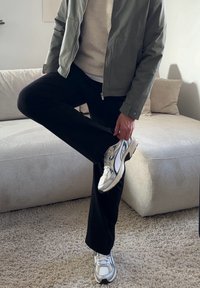 Personne portant une veste grise, un pull beige, un pantalon noir et des baskets blanches, debout sur une jambe à côté d'un canapé beige.