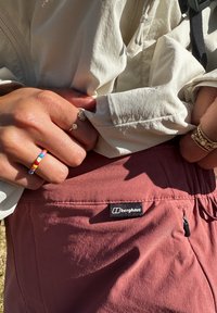 Bordeaux shorts med elastisk linning, med en glidelåslomme på siden. En beige jakke henger over livet. Berghaus-logoen er synlig.