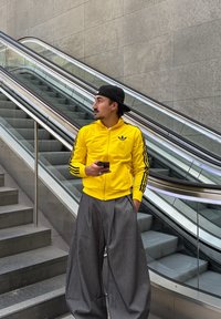 Sárga cipzáras dzseki fekete csíkokkal és logóval, szürke, bő szárú nadrággal párosítva. A személy okostelefont tart, és egy mozgólépcső közelében áll.