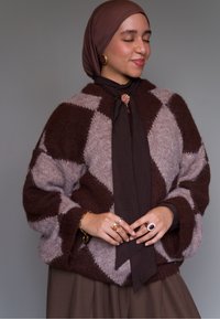 Pulover supradimensionat maro și bej cu model �în formă de diamant și textură moale. Poartă un fular maro închis și inele multicolore.