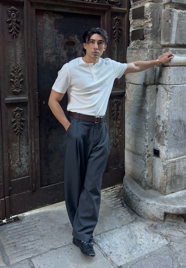 Hombre con camisa blanca de manga corta y pantalones oscuros apoyado contra una esquina de piedra cerca de una puerta de metal oscuro ornamentada sobre pavimento de piedra.