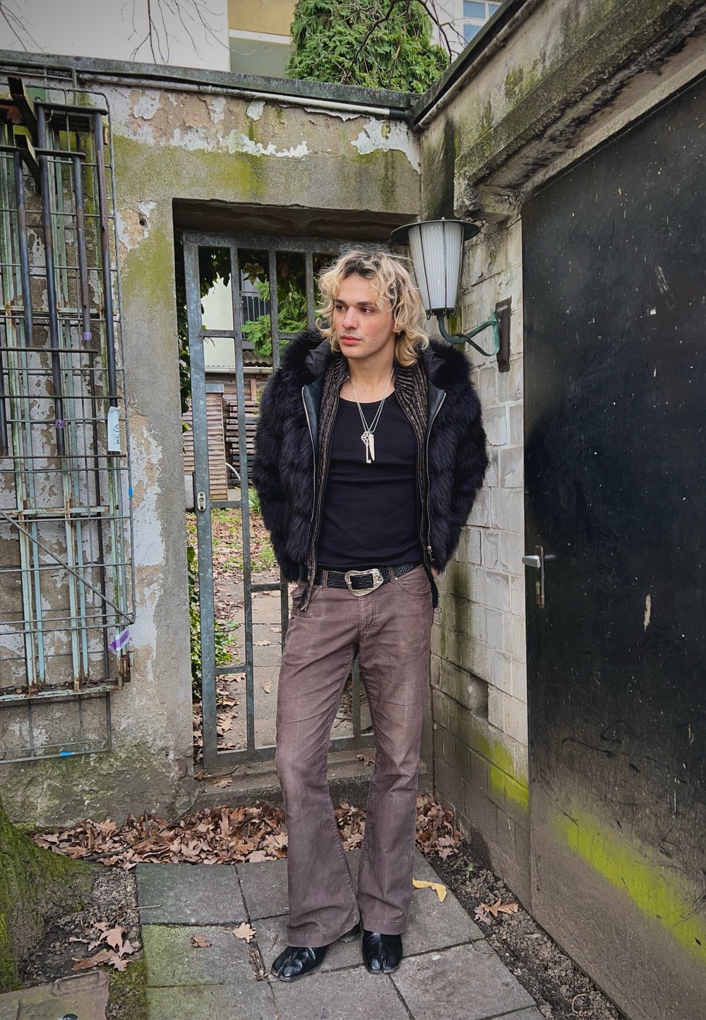 Μαύρο γελέκο από faux γούνινη ύφανση με δερμάτινη εσωτερική επένδυση, μαύρο τοπ με αθλητική εφαρμογή, καφέ φαρδιά παντελόνια και μαύρα παπούτσια; στυλιζαρισμένο με διαδοχικά κολιέ.