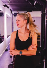 Black sports vest, slim-fit design; wristwatch with a round face and digital display; ponytail; background includes gym equipment and lighting.