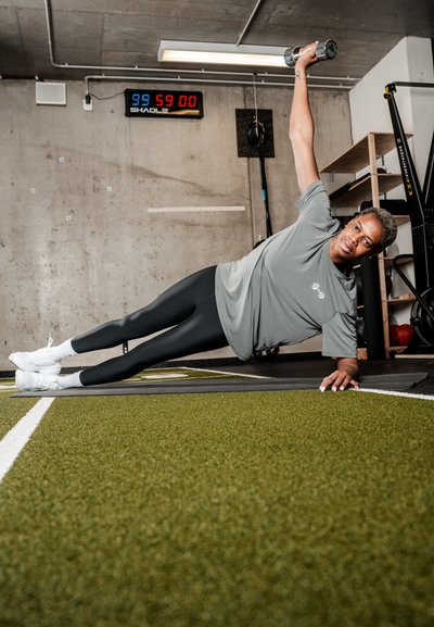 Persona realizando ejercicio de plancha lateral sobre una esterilla, sosteniendo una mancuerna por encima de la cabeza en un gimnasio con paredes de concreto y un temporizador visible.
