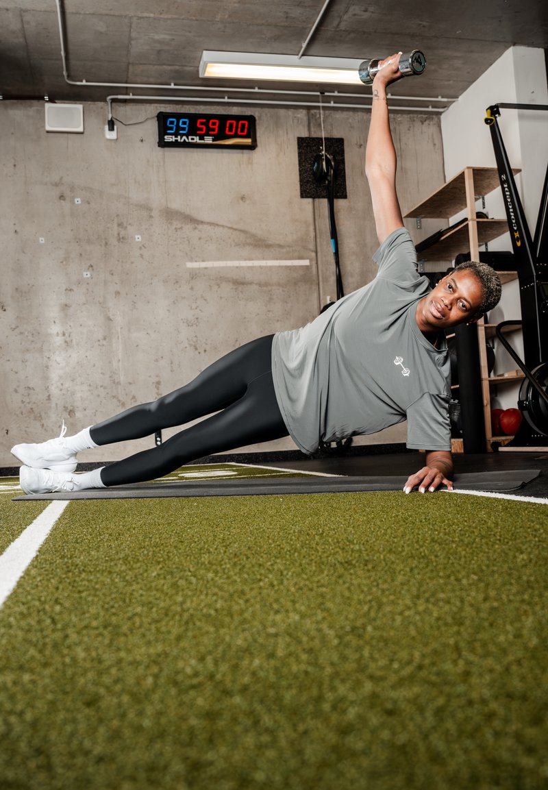 Persona che esegue un esercizio di plank laterale su un tappetino, tenendo un manubrio sollevato sopra la testa in una palestra con pareti in cemento e display del timer.