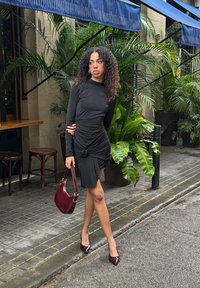 Fekete hosszú ujjú ruha, melyen begyűrt részletek, csomós díszítés és aszimmetrikus szegély található. Bordó táskával és magassarkú cipővel kiegészítve.