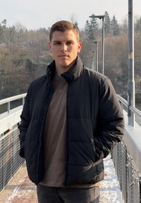 Fekete bélelt dzseki magas gallérral, barna ing fölött viselve. Egy hídon áll, szürke farmernadrágban, háttérben zöld növényzet és egy víztömeg látható.