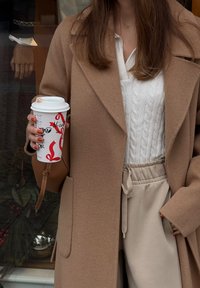 Manteau croisé camel porté sur un pull blanc à torsades, associé à un pantalon de jogging beige. Tenant une tasse de café ornée d'un ruban rouge festif.