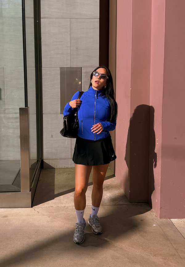Mujer con chaqueta azul y falda negra está de pie a la luz del sol, sosteniendo un bolso negro y lleva gafas de sol, zapatillas deportivas y calcetines blancos.