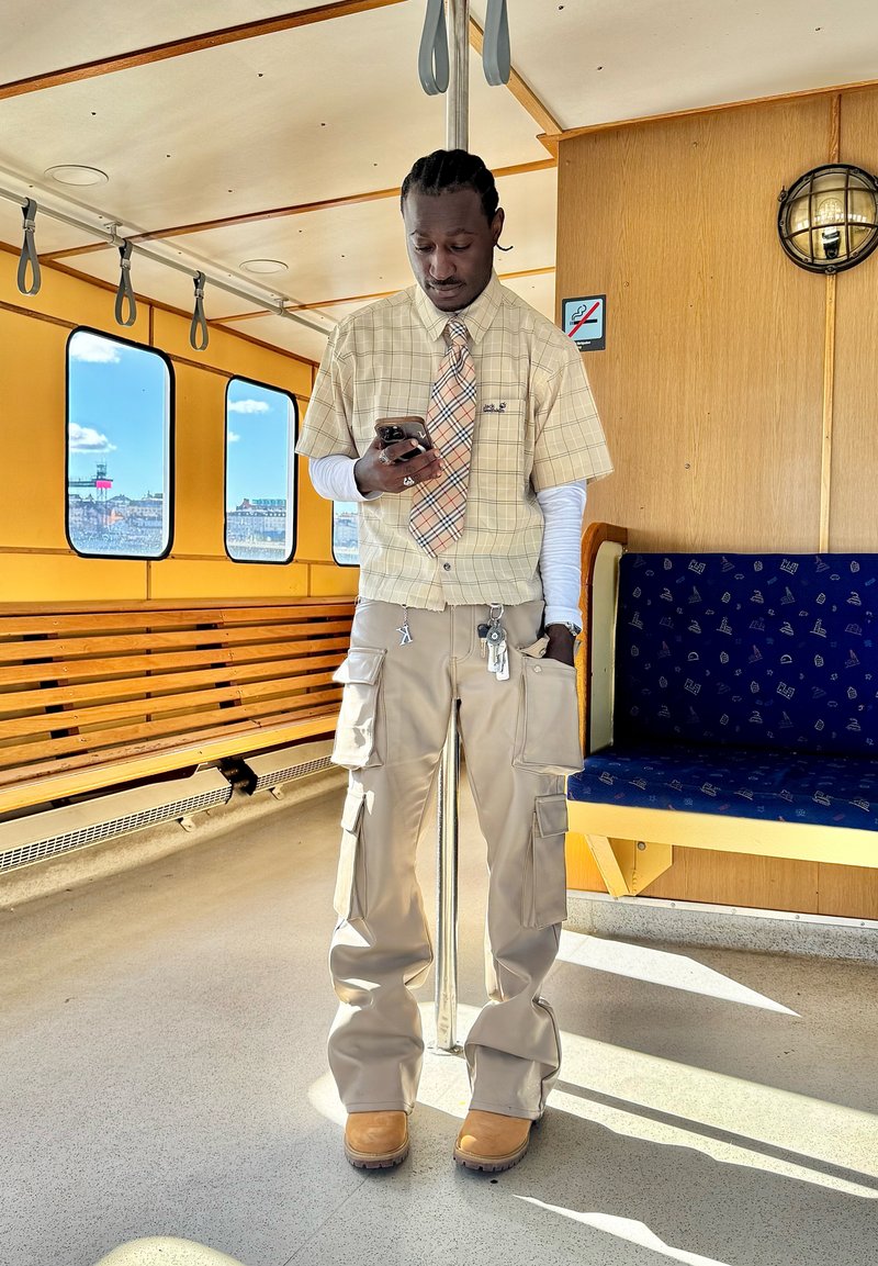 Homme portant un pantalon cargo beige, une chemise à manches courtes à carreaux et une cravate à motifs, debout à l'intérieur d'un véhicule de transport en commun jaune et bleu utilisant un smartphone.