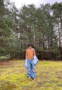 Giacca marrone con zip e dettagli blu, jeans in denim strappati oversize, e giacca in denim blu a strati, ambientati in una radura nel bosco.