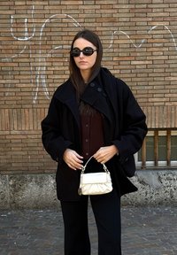 Femme en manteau noir et lunettes de soleil tenant un sac à main blanc, debout devant un mur de briques couvert de graffiti.