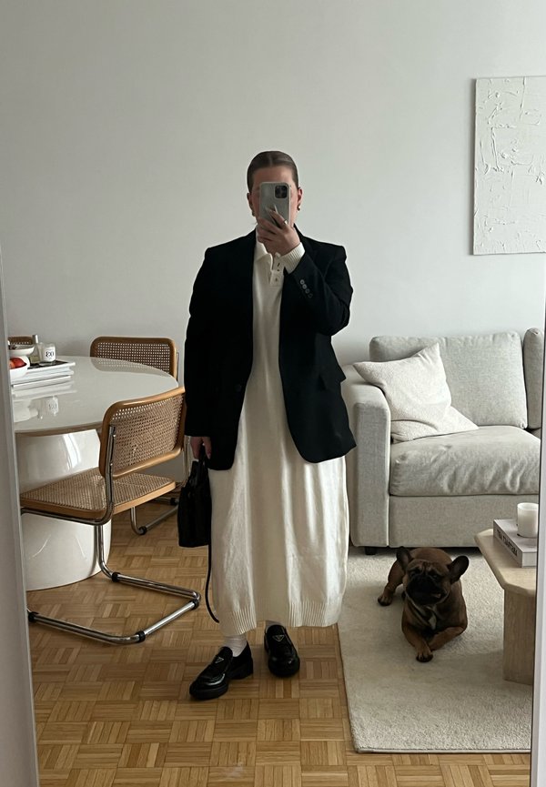 Eine Person trägt ein schwarzes Blazer über einem weißen Strickkleid, schwarze Mokassins und hält eine kleine Handtasche. Ein brauner Hund liegt auf dem Boden.