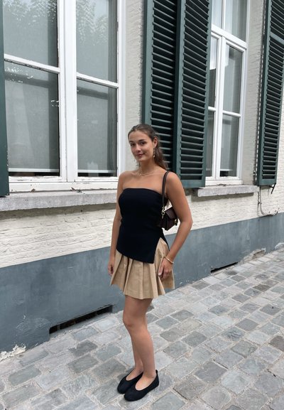 Top sin tirantes de franjas negras combinado con una falda plisada beige. Bailarinas negras sencillas y un pequeño bolso oscuro. Fondo de calle de adoquines.