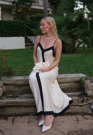 Vestido color crema con un diseño geométrico negro, tejido texturizado y una gran abertura, combinado con tacones blancos de punta, sosteniendo un ramo de flores.