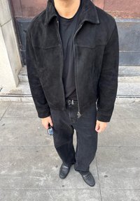Jachetă din suede neagră cu fermoar, purtată peste un tricou negru. Pantaloni din denim negri și pantofi texturați negri. Accesoriile includ ochelari de soare.