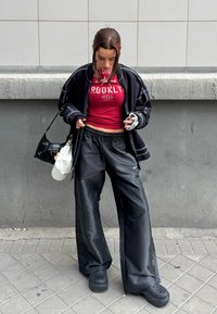 Jeune femme en pantalon large noir, crop top rouge et veste noire, regardant vers le bas, debout sur un trottoir urbain contre un mur gris.