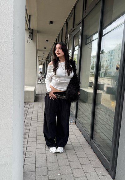 Top blanco de manga larga con diseño drapeado, pantalones negros sueltos y un accesorio de piel sintética negra. Combinado con calzado blanco, de pie sobre pavimento de baldosas.