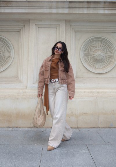 Mujer con cabello largo y oscuro, lleva gafas de sol grandes, chaqueta marrón, camiseta de color óxido, pantalones anchos y claros, zapatos beige, sosteniendo un bolso beige, caminando junto a una pared ornamentada.