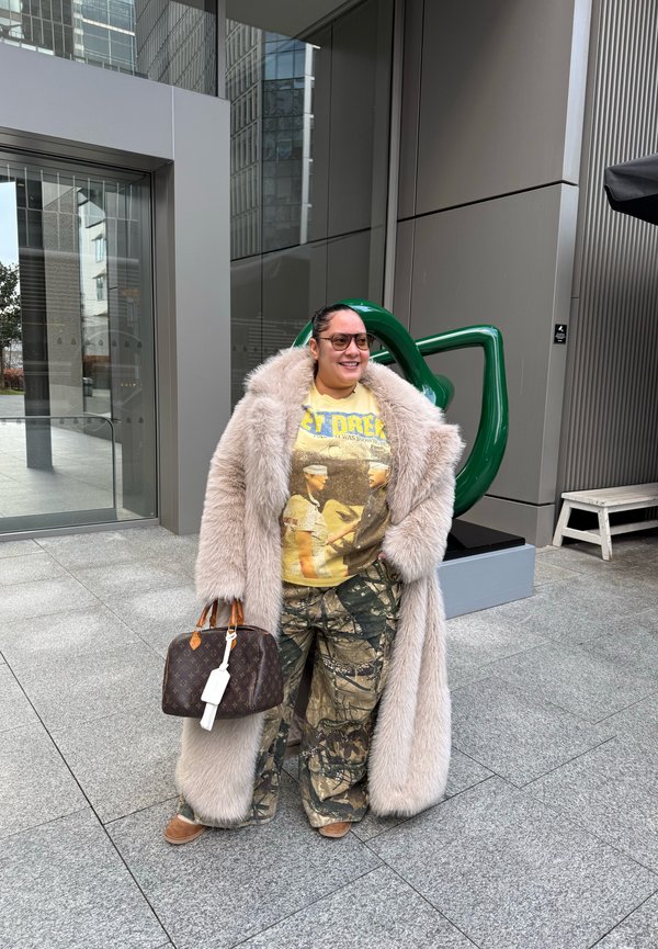 Persoană care poartă o haină de blană bej peste un tricou galben Star Trek și pantaloni de camuflaj, ținând o geantă maro de designer în aer liber, lângă clădiri moderne.