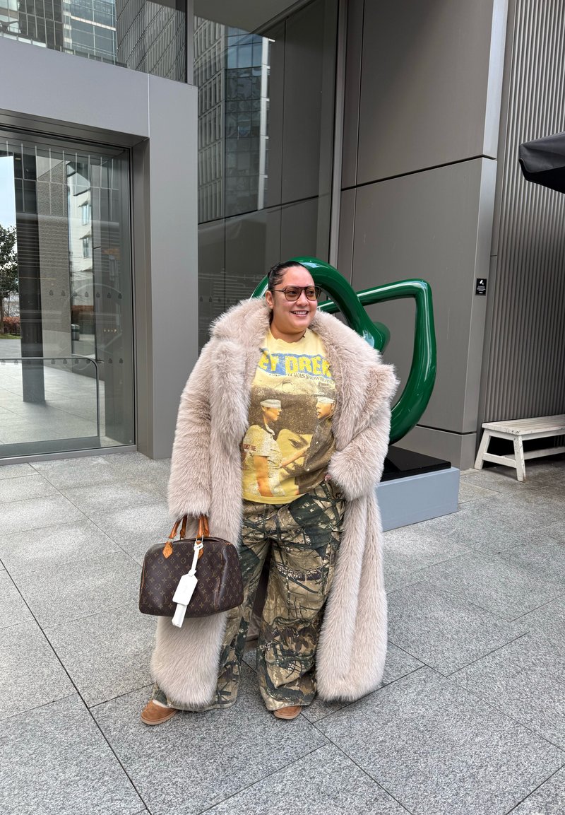 Personne portant un manteau en fausse fourrure beige par-dessus un t-shirt jaune Star Trek et un pantalon camouflage, tenant un sac à main de créateur brun à l'extérieur près de bâtiments modernes.