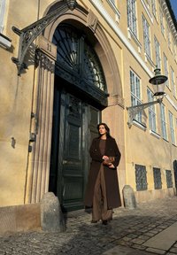 Καφέ παλτό, ταιριαστό σύνολο, στέκεται δίπλα σε μια διακοσμημένη πράσινη πόρτα με μαύρο υλικό. Διαδρομή από πέτρες, υφή τοίχου και φανοστάτης ορατά.