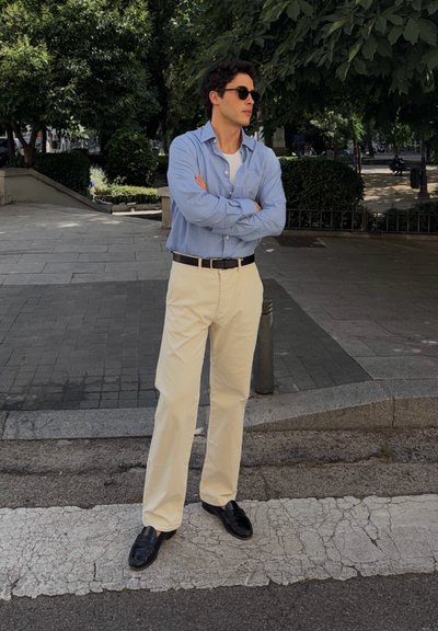 Hombre con gafas de sol, camisa azul, pantalones beige y zapatos negros, de pie con los brazos cruzados en una calle pavimentada cerca de árboles y arbustos.