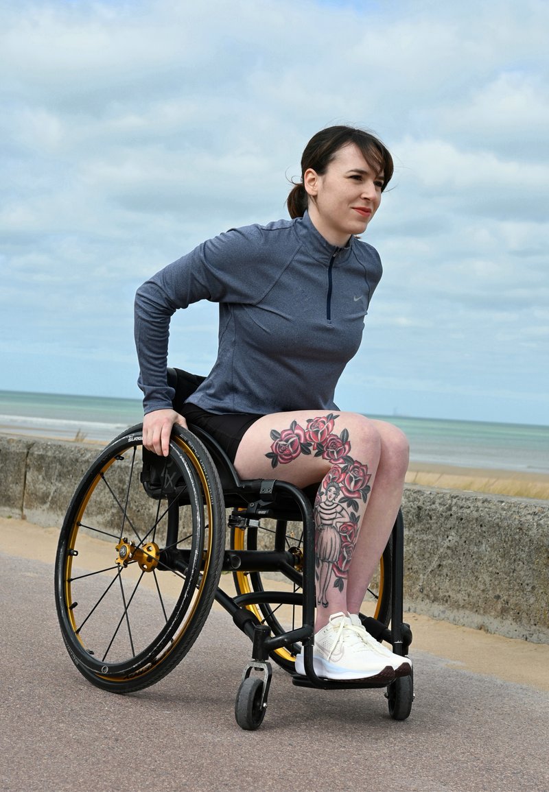 Naine ratastoolis, kelle jalal on lille- ja figuuritatueringud, riides spordiriietesse, liikumas väljas mereäärse müüri lähedal pilvise taeva all.