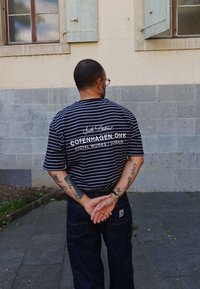 Mężczyzna z tatuażami, ubrany w czarną, paskowaną koszulę z napisem "Copenhagen DNK", stojący z dłońmi złożonymi za plecami przy kamiennej ścianie.
