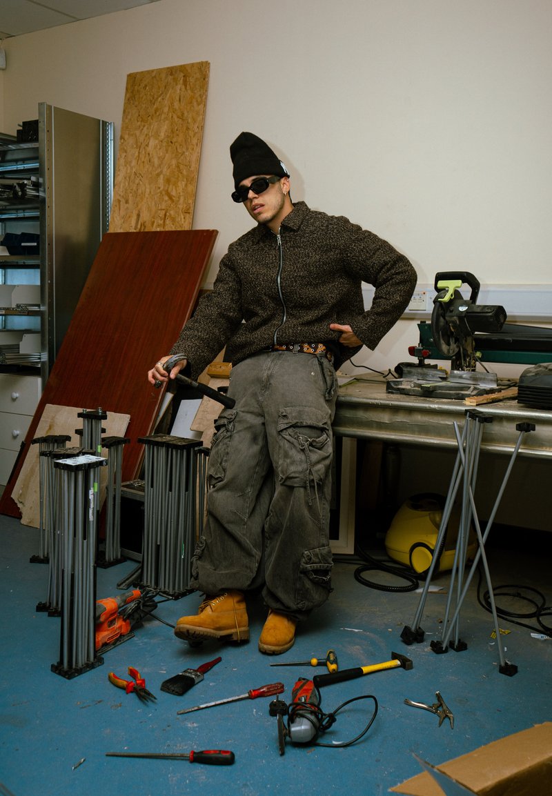 A person dressed in a brown textured zip-up jumper, grey cargo trousers, and yellow work boots, positioned amidst various tools on a blue floor.