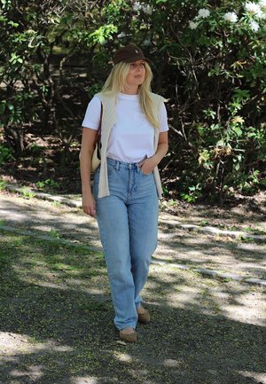 Conjunto informal que incluye una camiseta blanca, un chaleco beige, jeans claros de cintura alta y una gorra marrón. Texturas suaves y corte relajado. Fondo natural.