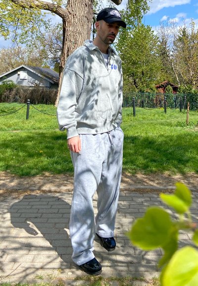 Hombre con sudadera y pantalones deportivos grises, con zapatos negros y gorra, de pie en un camino pavimentado con césped verde y árboles al fondo.