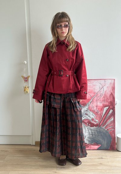 Joven con chaqueta roja con cinturón, falda larga de cuadros y gafas de sol, de pie en el interior cerca de una pintura de unicornio en el suelo.