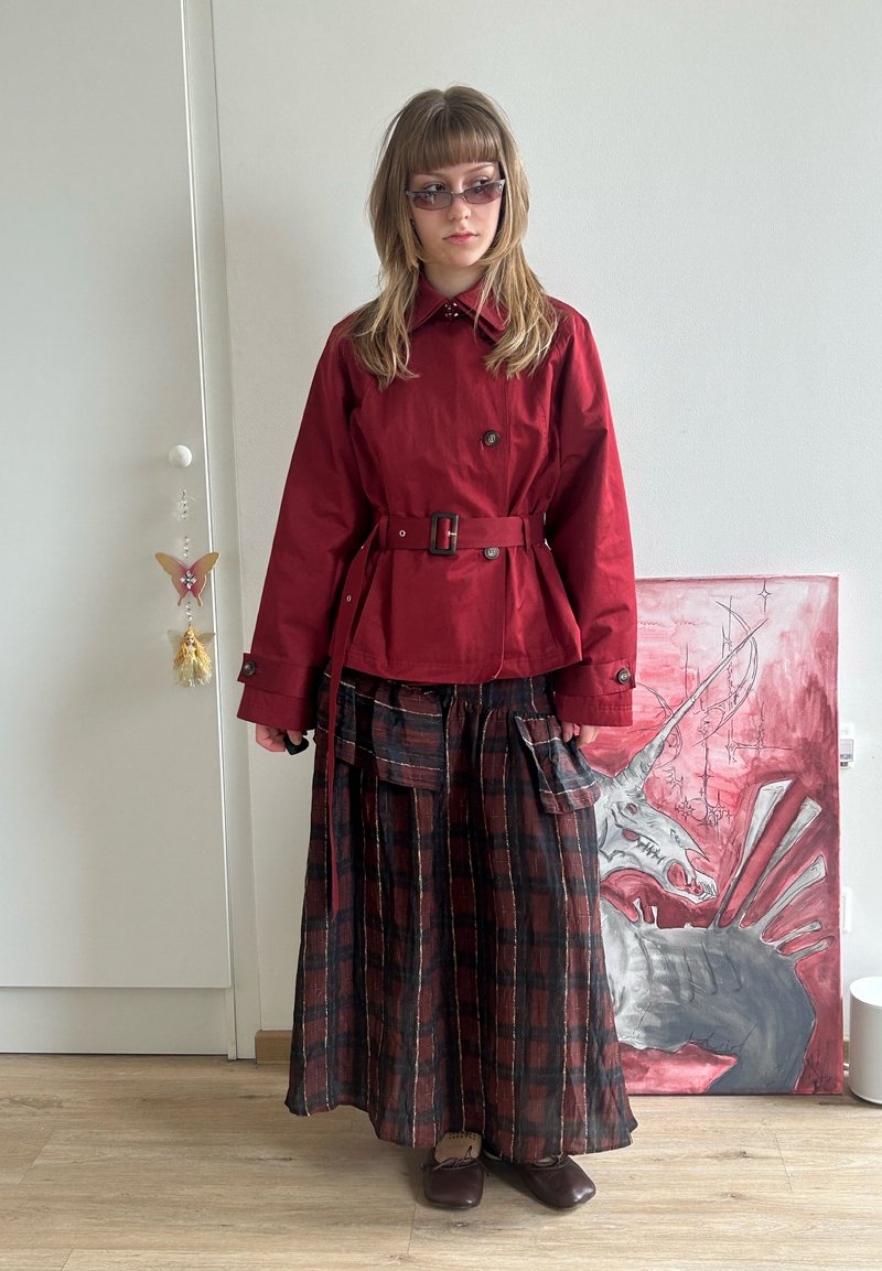 O tânără care poartă o jachetă roșie cu curea, o fustă lungă în carouri și ochelari de soare stă în interior, lângă o pictură cu un unicorn pe podea.