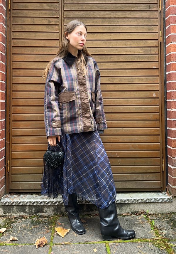 Gepolsterte Karo-Jacke in Braun und Marineblau, kombiniert mit einem karierten Rock in Marineblau. Das Model hält eine schwarze herzförmige Perlenhandtasche. Schwarze kniehohe Stiefel.