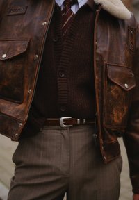 Brun skinnjakke med pelskrage, brun strikkejakke, mønstret slips, brun belte med sølvspenne, og grå bukser med kammerspor.