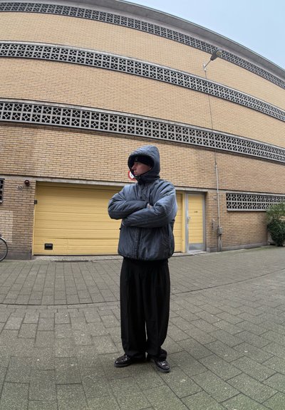 Chaqueta oversized negra, pantalones negros y zapatos, de pie en una calle pavimentada. Puerta de garage amarilla al fondo; pared de ladrillo texturizada.