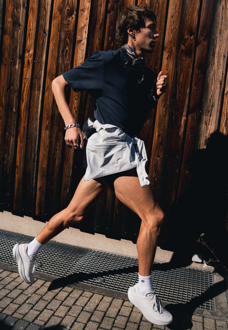 Person som løper utendørs iført svart skjorte, hvit jakke bundet rundt livet, hvite joggesko og mønstret halskjede foran en trevegg.