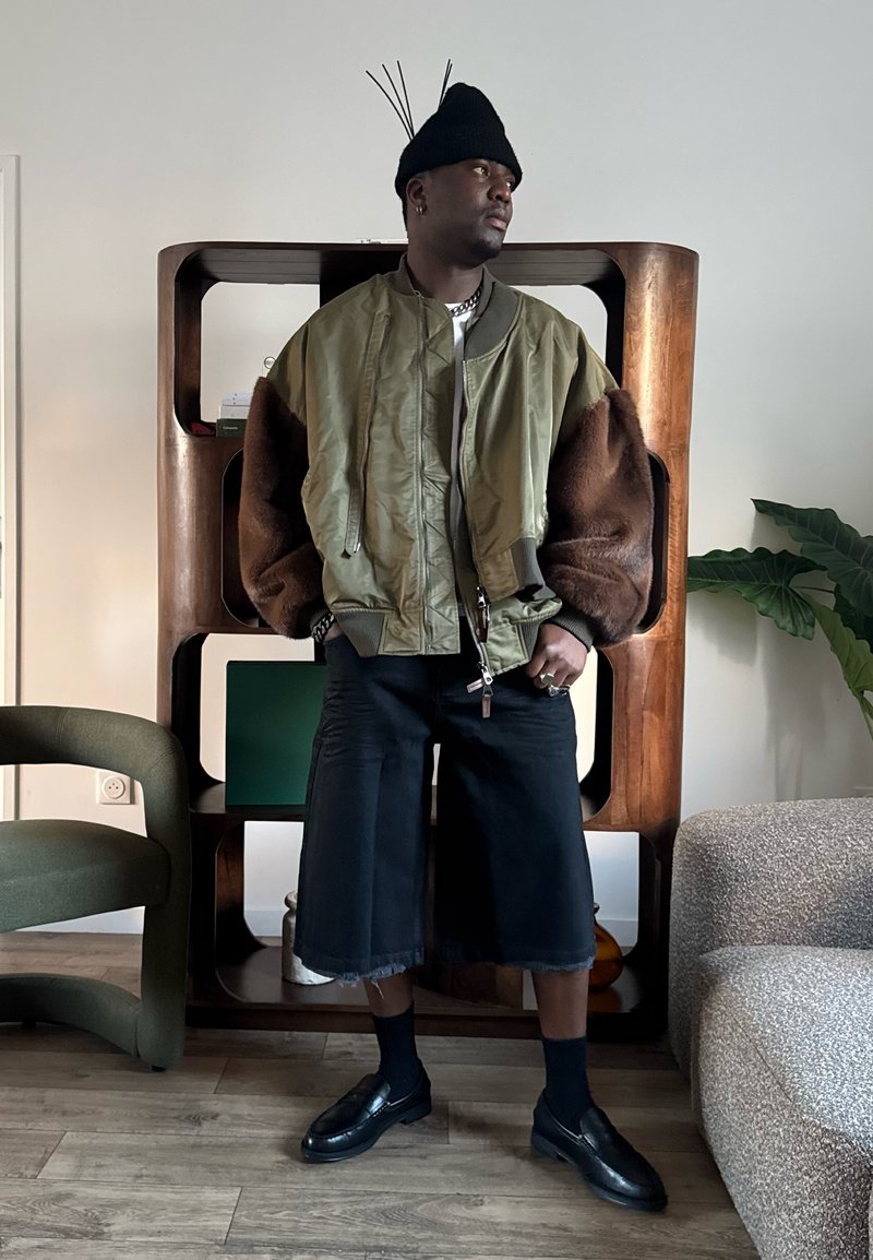 Homme portant un bonnet noir avec des bâtons, une veste bomber verte avec des manches en fourrure marron, un short large noir et des mocassins noirs dans un salon moderne.