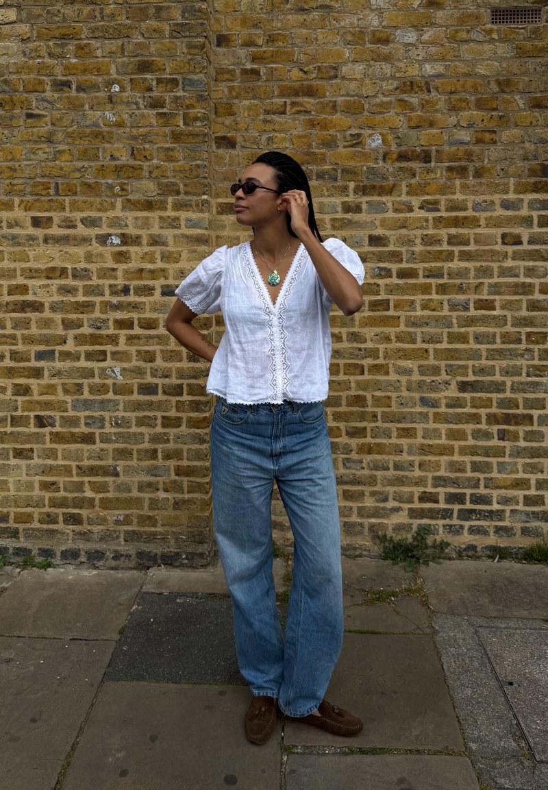 Femme avec des lunettes de soleil portant un chemisier en dentelle blanche, un jean bleu et des chaussures marron, debout contre un mur de briques jaunes, ajustant sa boucle d’oreille.
