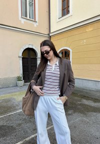 Rudas švarkas ant dryžuoto polo marškinėlių, baltos laisvo kirpimo kelnės ir ruda petneša. Akiniai nuo saulės suteikia kontrastą aprangai.
