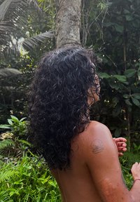 Personne aux cheveux bouclés et foncés avec un tatouage de coquillage sur l'épaule, debout torse nu dans une forêt verdoyante, face à droite avec les mains légèrement levées.