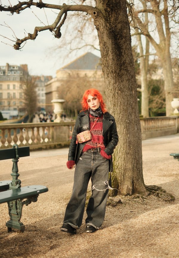 Pessoa a usar uma jaqueta de couro preta, um suéter vermelho felpudo, jeans largos e desgastados, e sapatos de plataforma, com cabelo laranja e uma bolsa estampada.