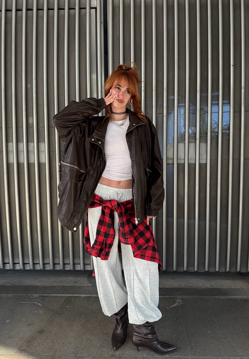 Jachetă oversized din piele ecologică neagră purtată peste un top scurt alb, pantaloni de trening gri și o cămașă în carouri roșu și negru legată în talie. Botine maro până la glezne.