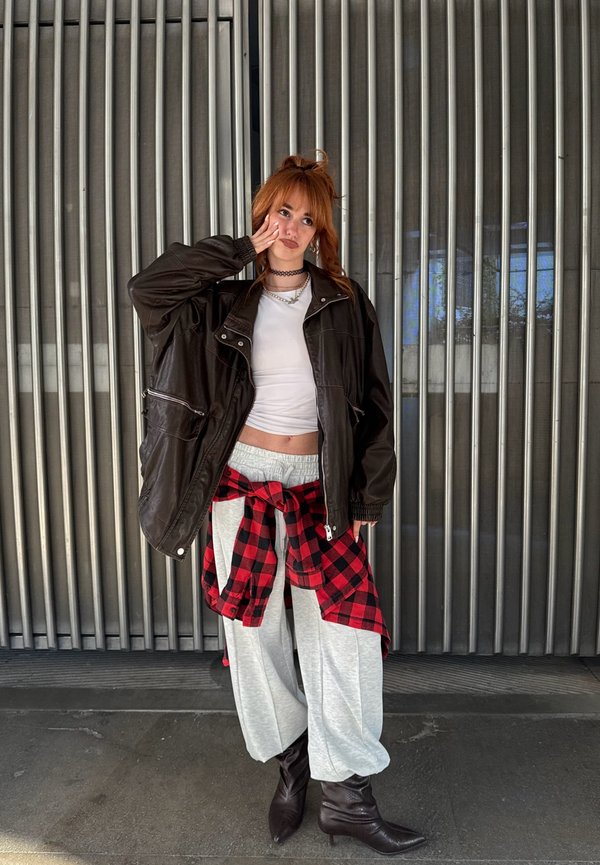 Överdimensionerad svart imitationsläderjacka över en vit crop top, gråa sweatpants och en röd och svart rutig skjorta knuten i midjan. Bruna ankelboots.