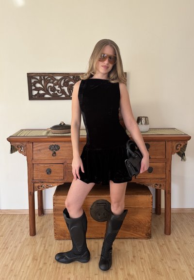 Vestido sin mangas de terciopelo negro con falda acampanada, combinado con botas negras de vinilo hasta la rodilla y un pequeño bolso negro.